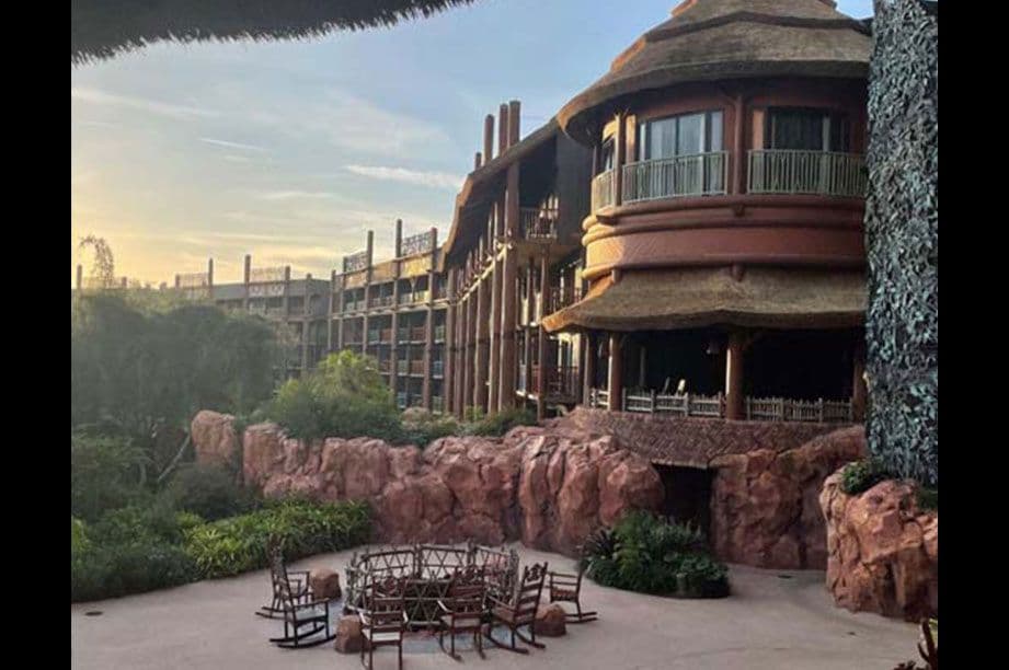 Curved multi-story Disney resort building with natural stone accents and thatched roof elements, featuring outdoor seating area with red rock formations.