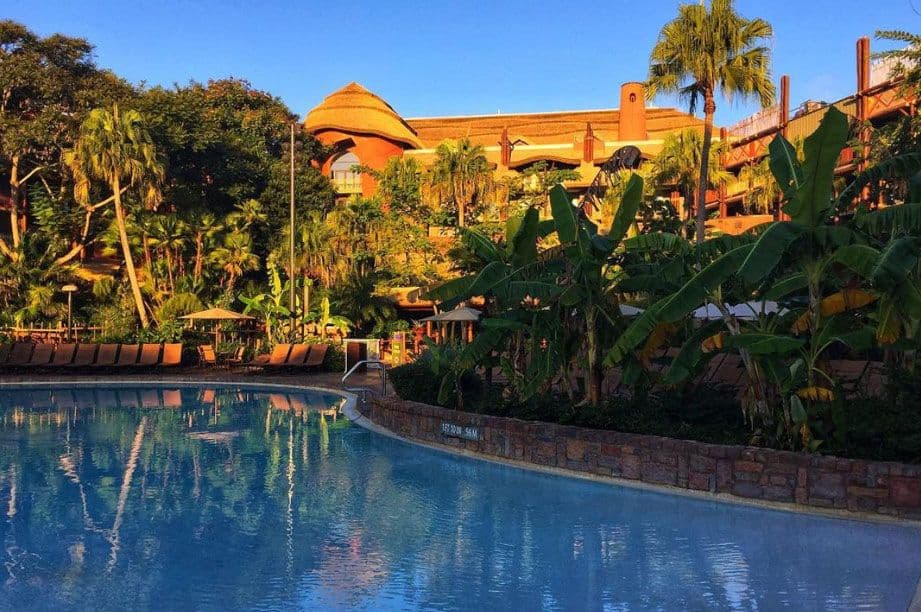 Resort pool surrounded by lush tropical landscaping with palm trees and banana plants, featuring colorful buildings in the background under a clear blue sky.