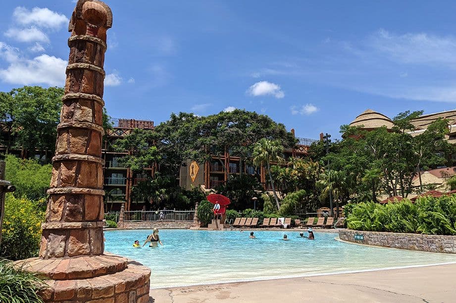 Stone pillar beside resort pool with guests swimming, surrounded by lush tropical landscaping and Disney resort buildings