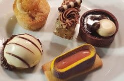 Five elegant desserts arranged on white plate: chocolate-covered treats, small cake, and pastries with various toppings and fillings.