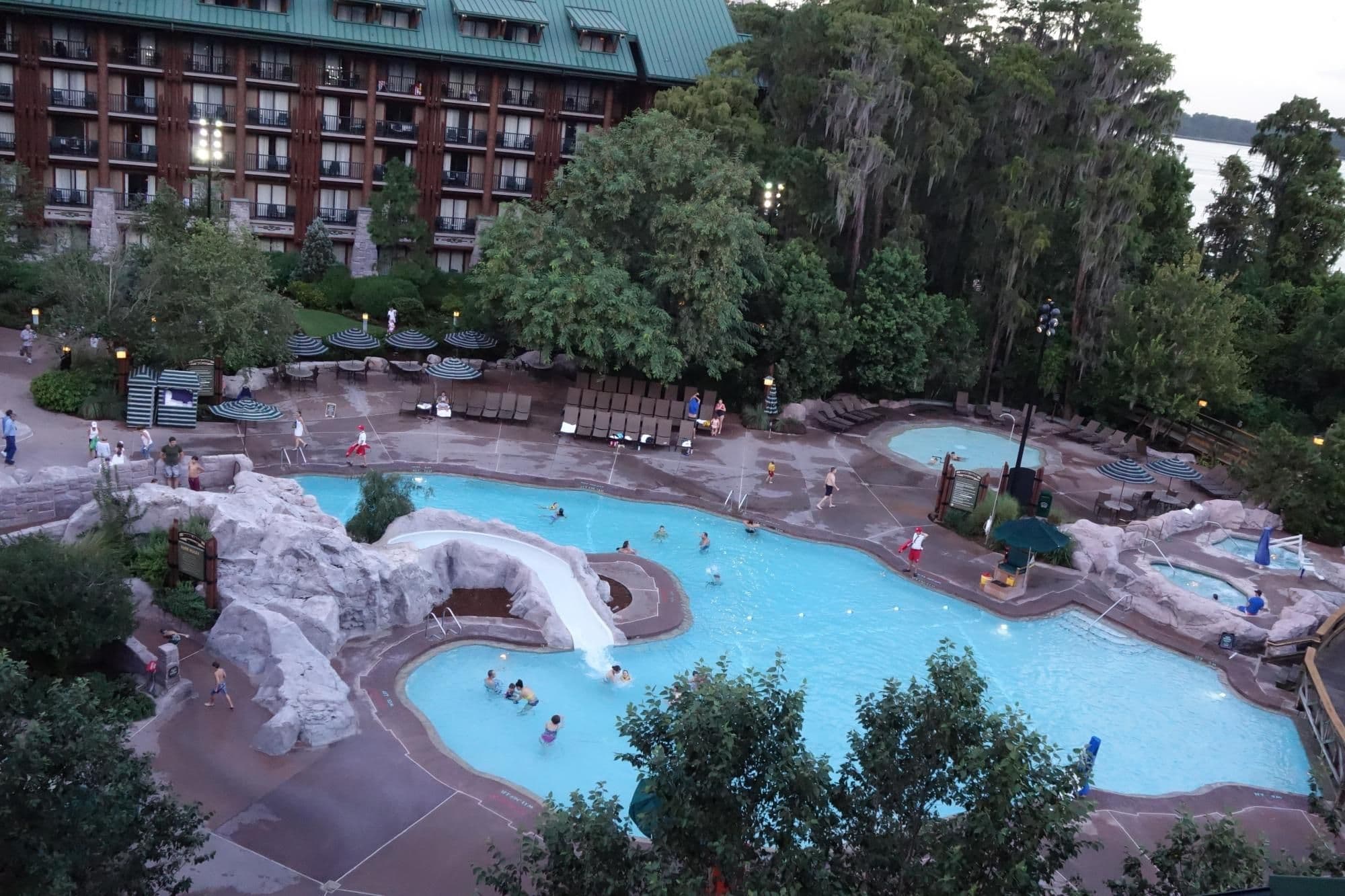 Curved swimming pool with hot tub at Disney resort, surrounded by lush trees and multi-story lodge building at dusk