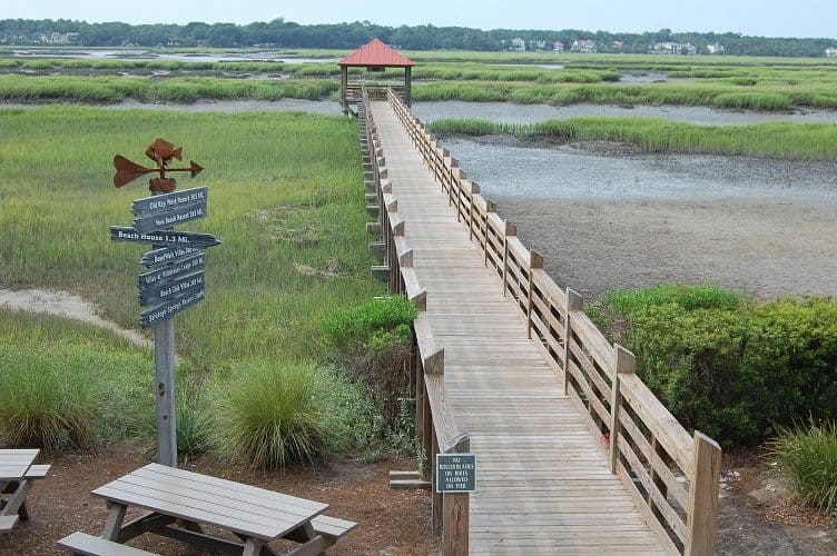 Wooden boardwalk with railings extends over salt marsh wetlands toward observation platform with red roof, directional signs visible.