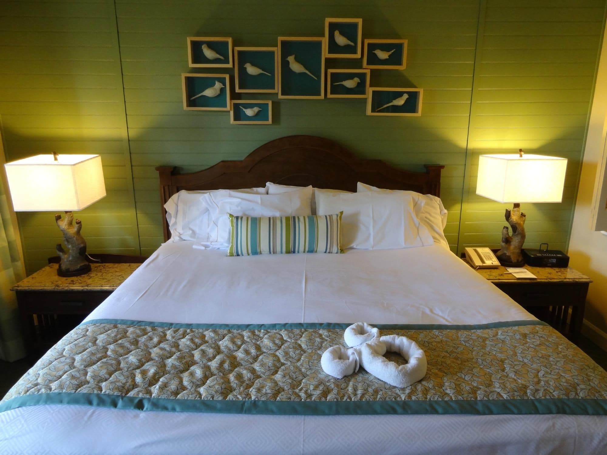 Hotel bedroom with white bedding, wooden headboard, bird artwork wall display, matching bedside lamps, and decorative towel swans on bed.