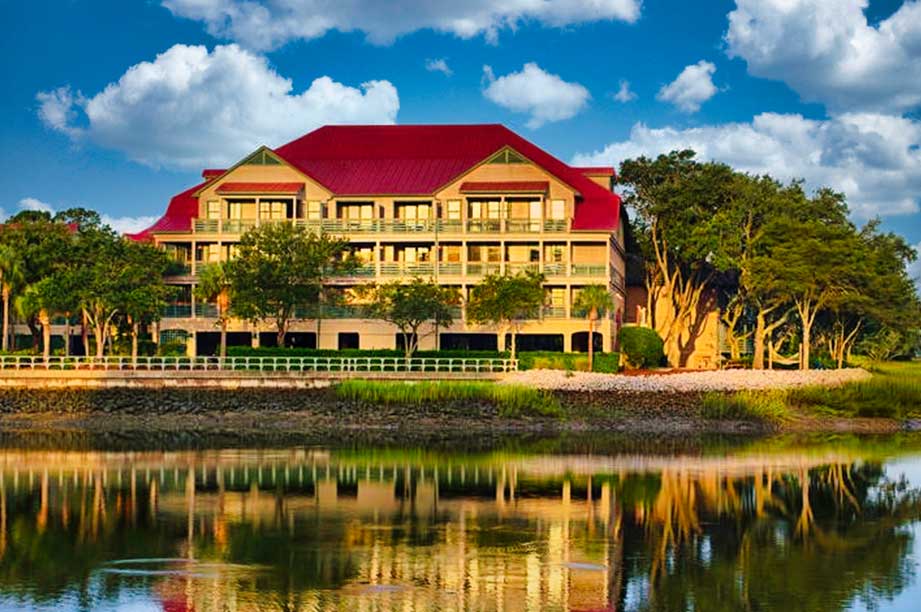 Waterfront Disney Vacation Club resort with red roof and yellow siding reflected in calm lake under blue sky with white clouds