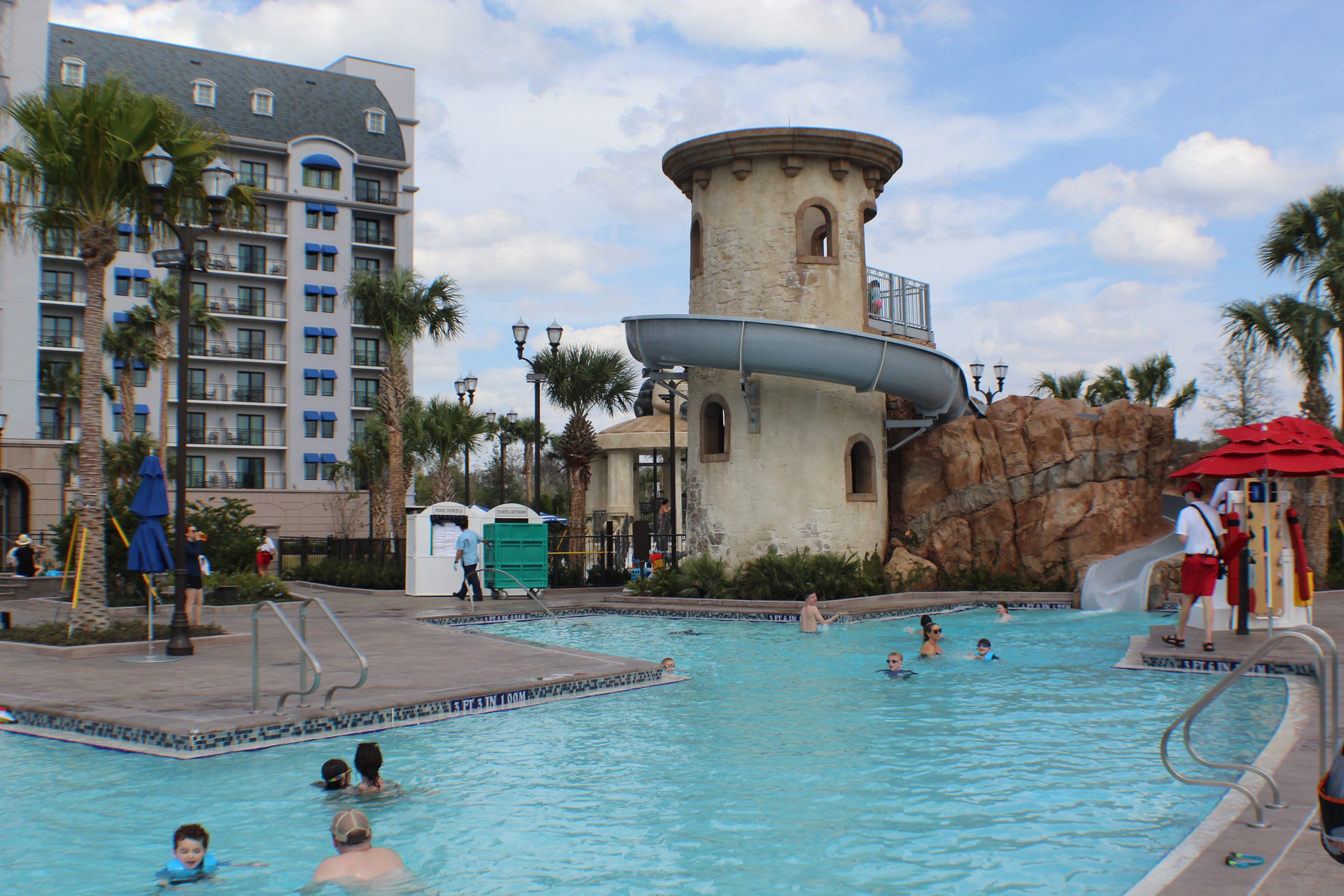 Pool area with castle-themed water slide tower, palm trees, and resort building in background under cloudy blue sky.
