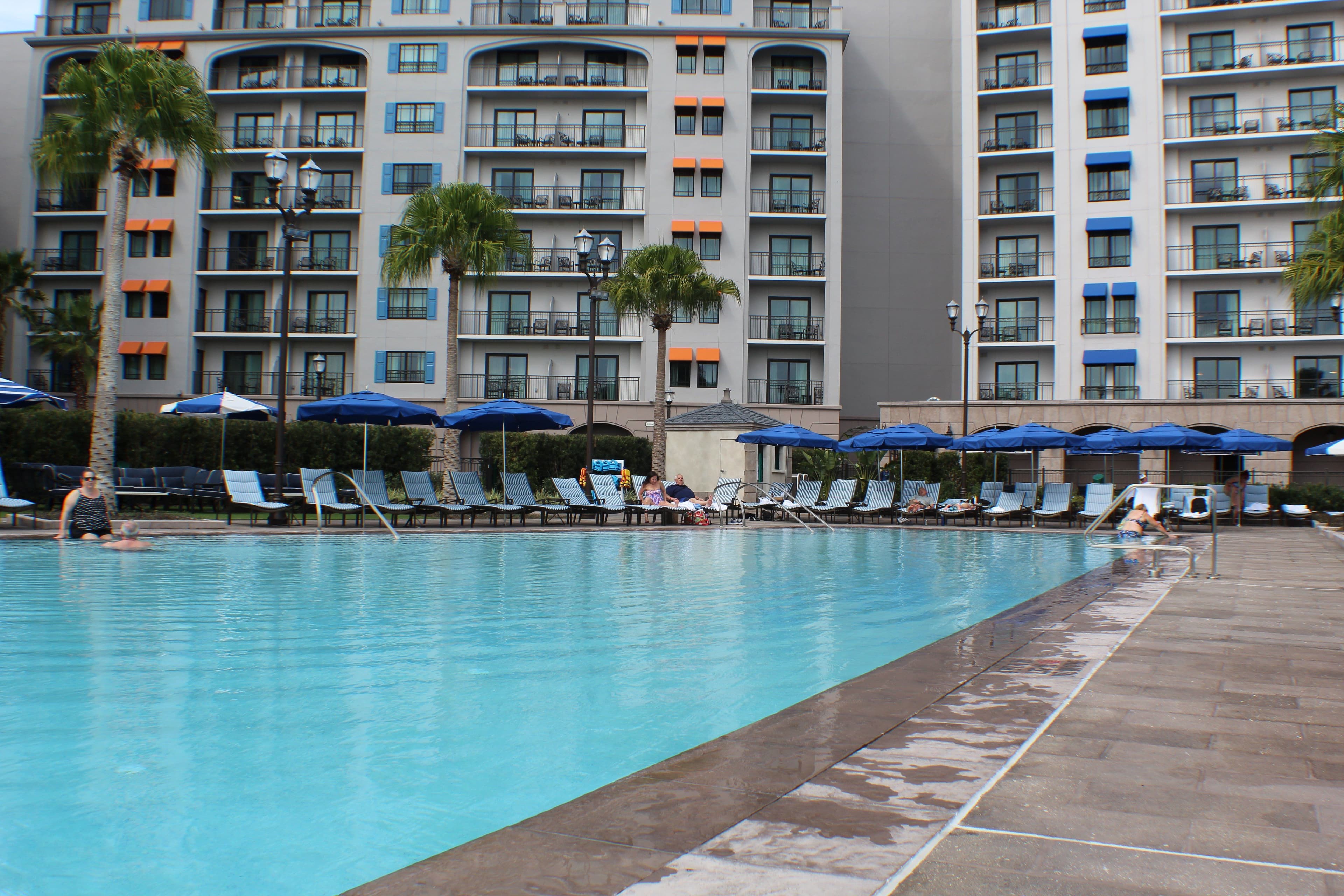 Large resort pool with blue umbrellas and lounge chairs, surrounded by multi-story vacation buildings with balconies and palm trees.