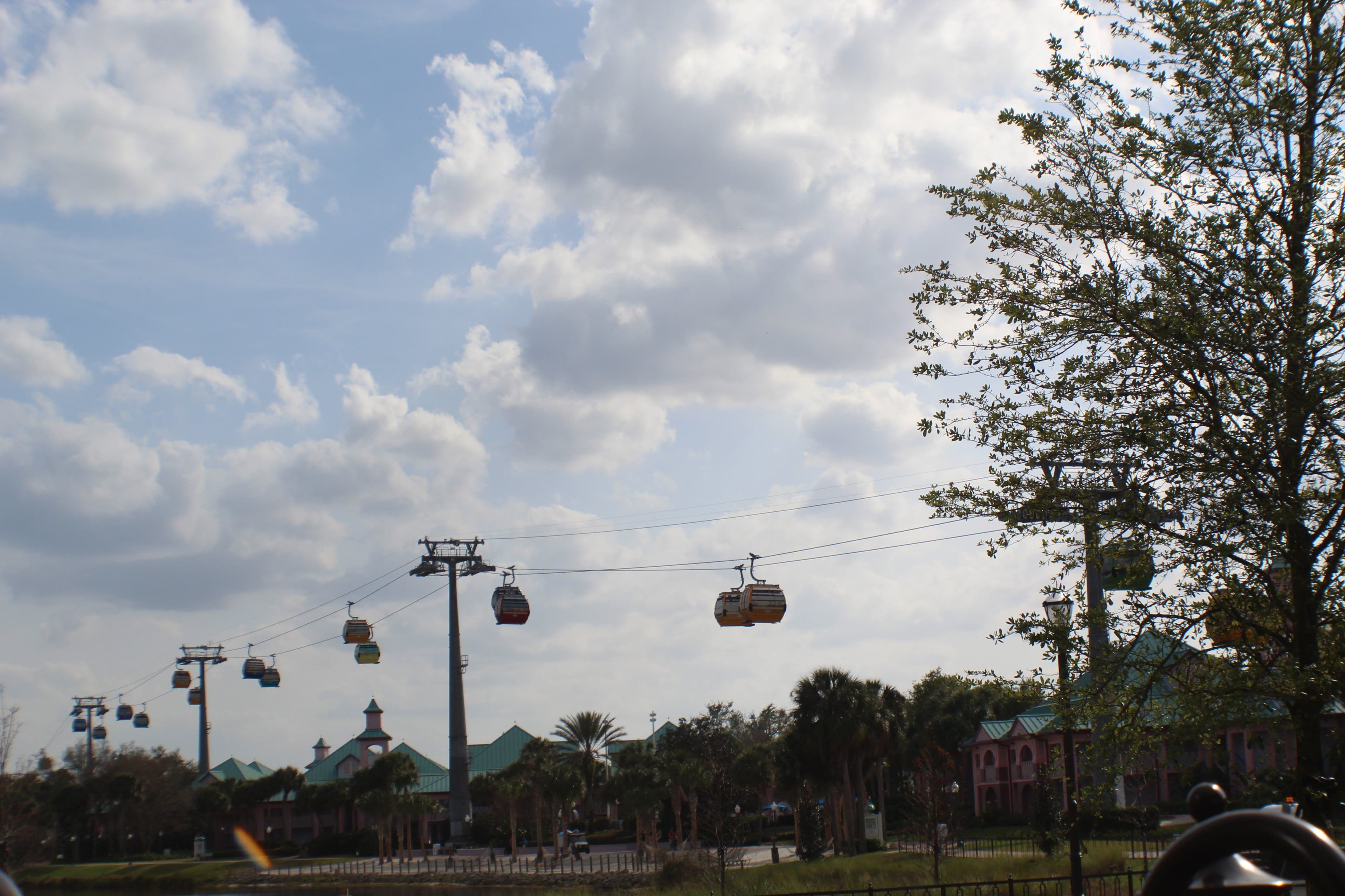 Disney Skyliner gondolas suspended on cables above resort buildings with palm trees and cloudy sky at Disney World.