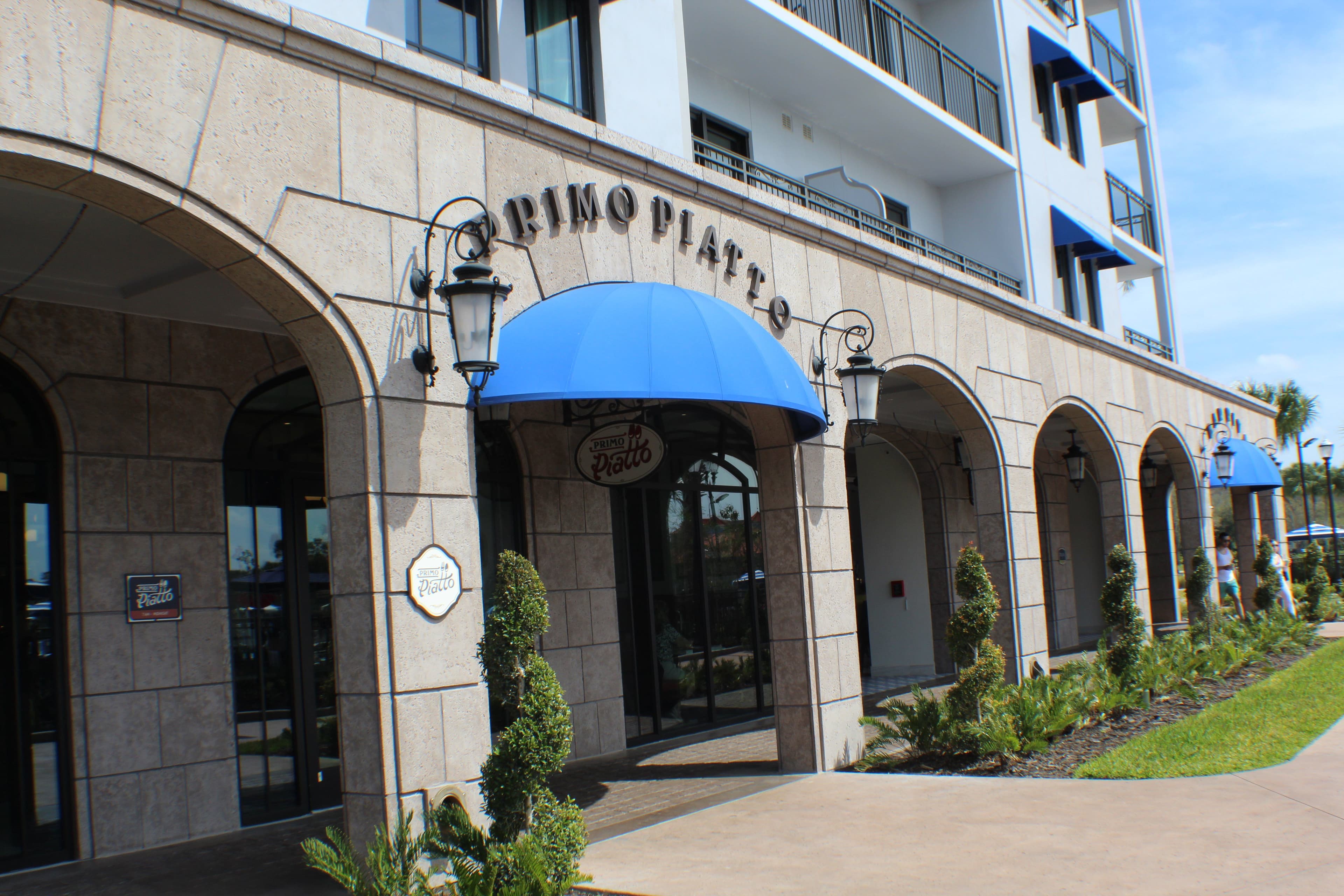 Stone archway entrance to Primo Piatto restaurant with blue awning, lantern-style lighting, and landscaped walkway at Disney resort.