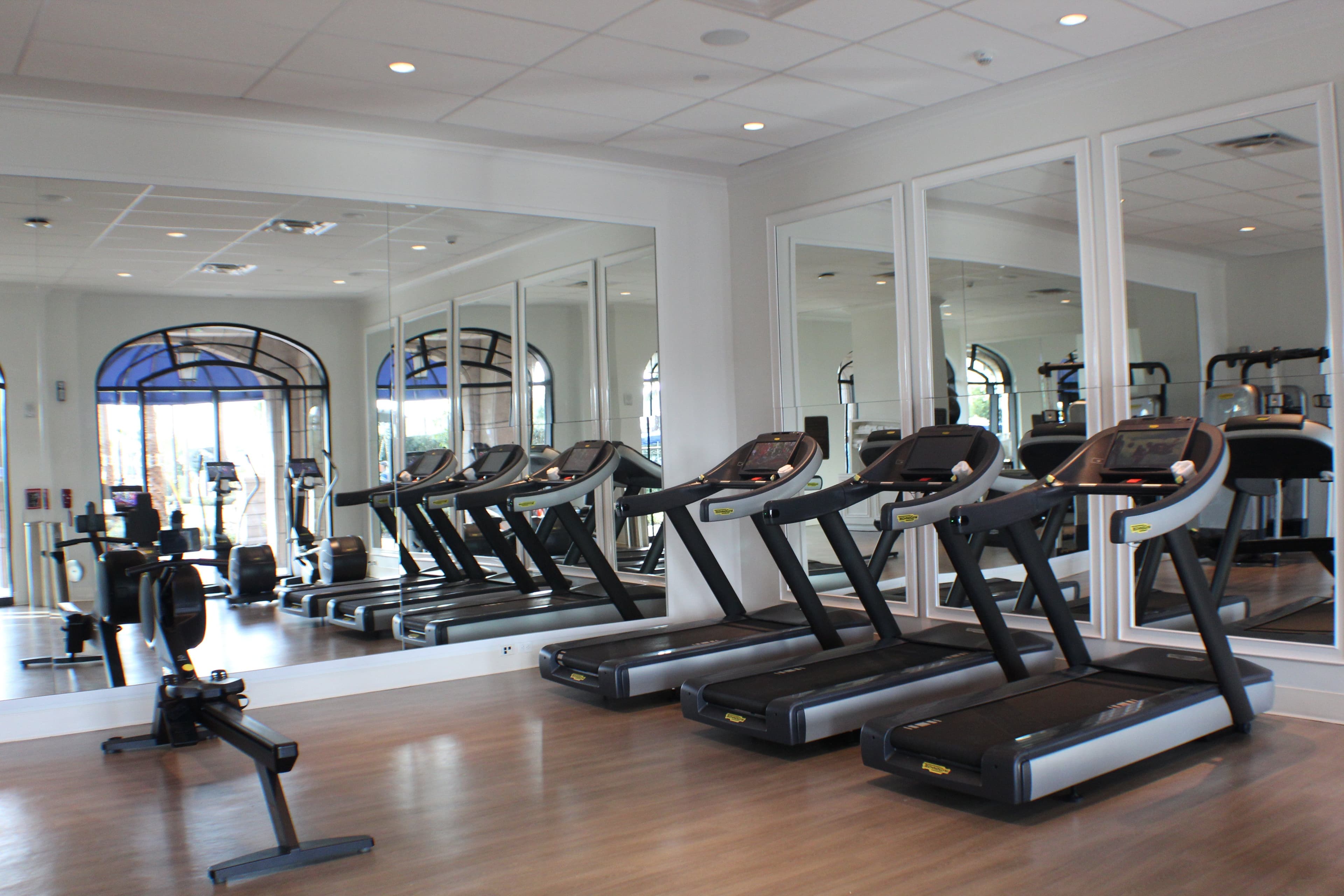Modern fitness center with row of treadmills, mirrored walls, arched windows, and exercise equipment on polished floors.