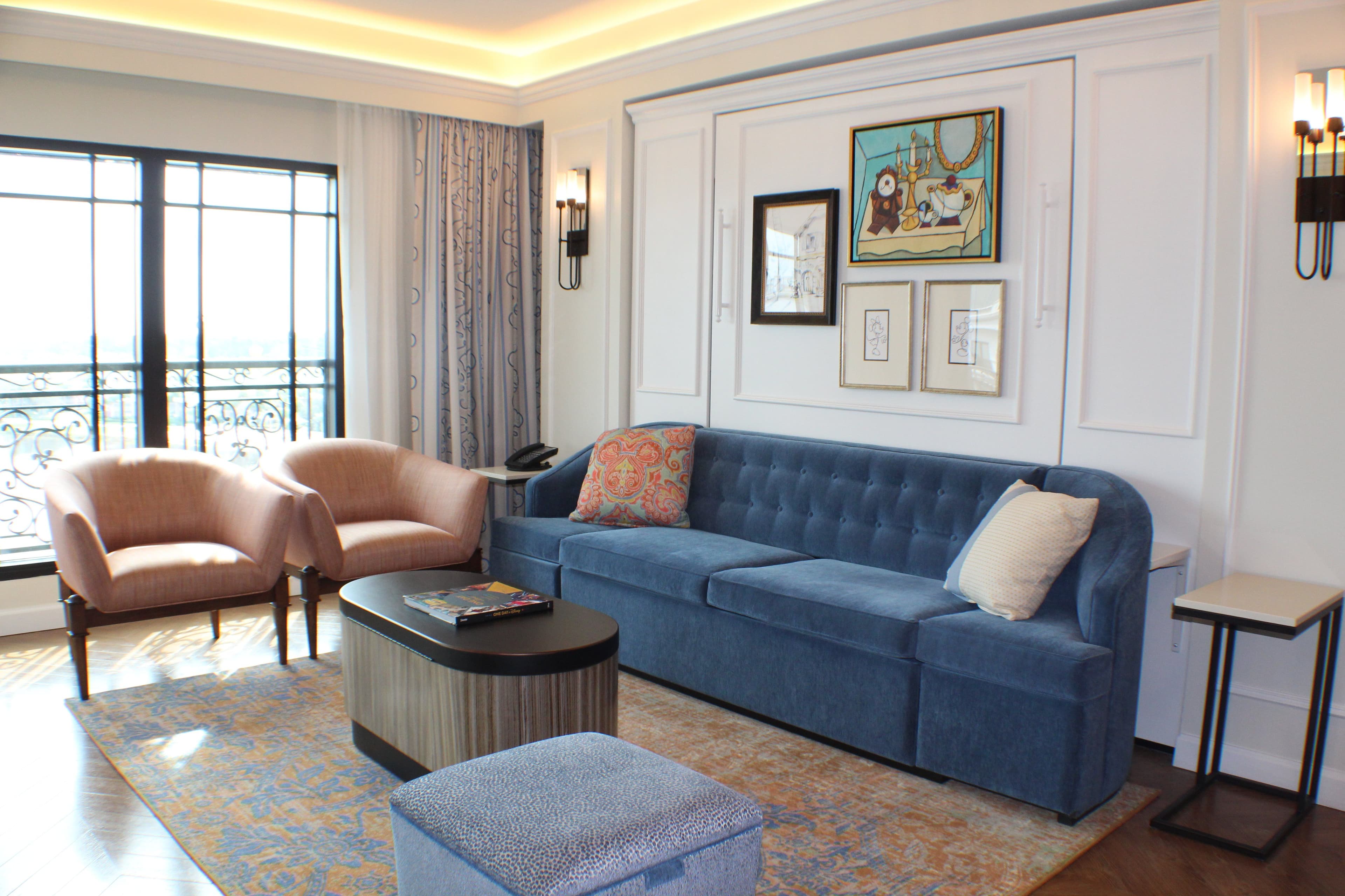 Elegant hotel suite living room with blue sofa, peach armchairs, coffee table, and French doors leading to balcony.