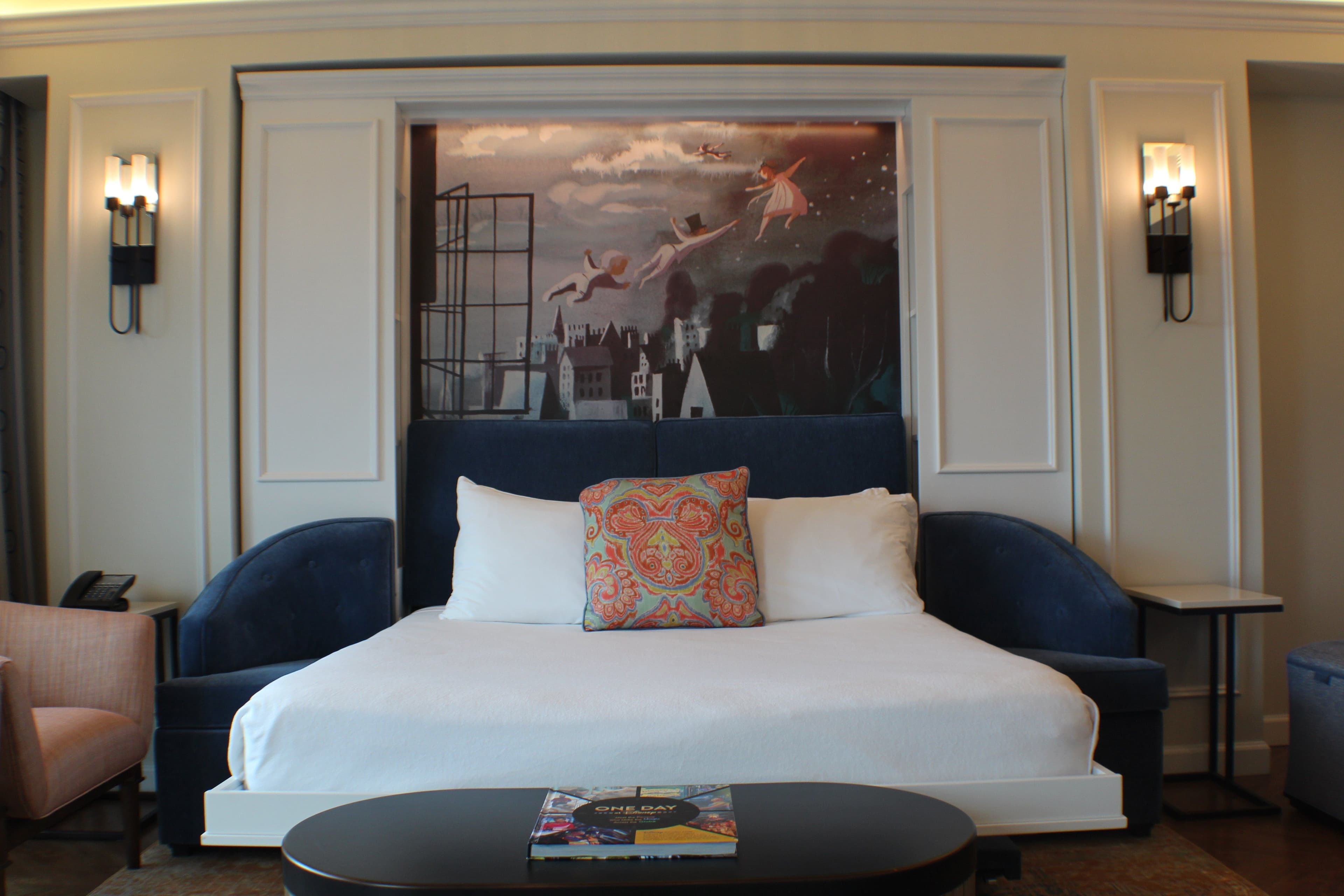 Elegant hotel room with white bed, navy upholstered headboard, decorative pillow, nautical artwork, and wall sconces in warm lighting.