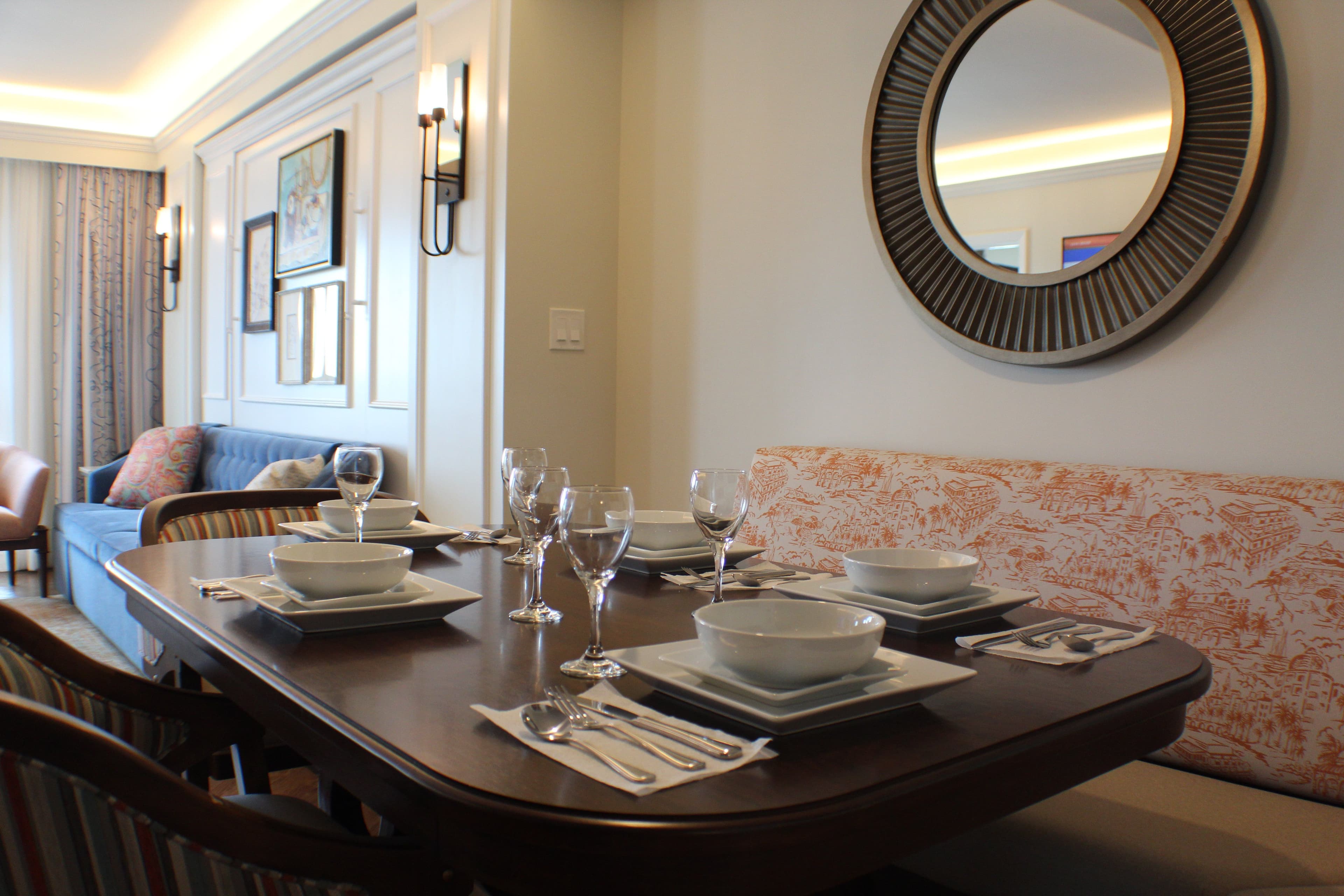 Elegant dining area with dark wood table set for four, decorative round mirror, and coral-patterned banquette seating in modern resort suite.