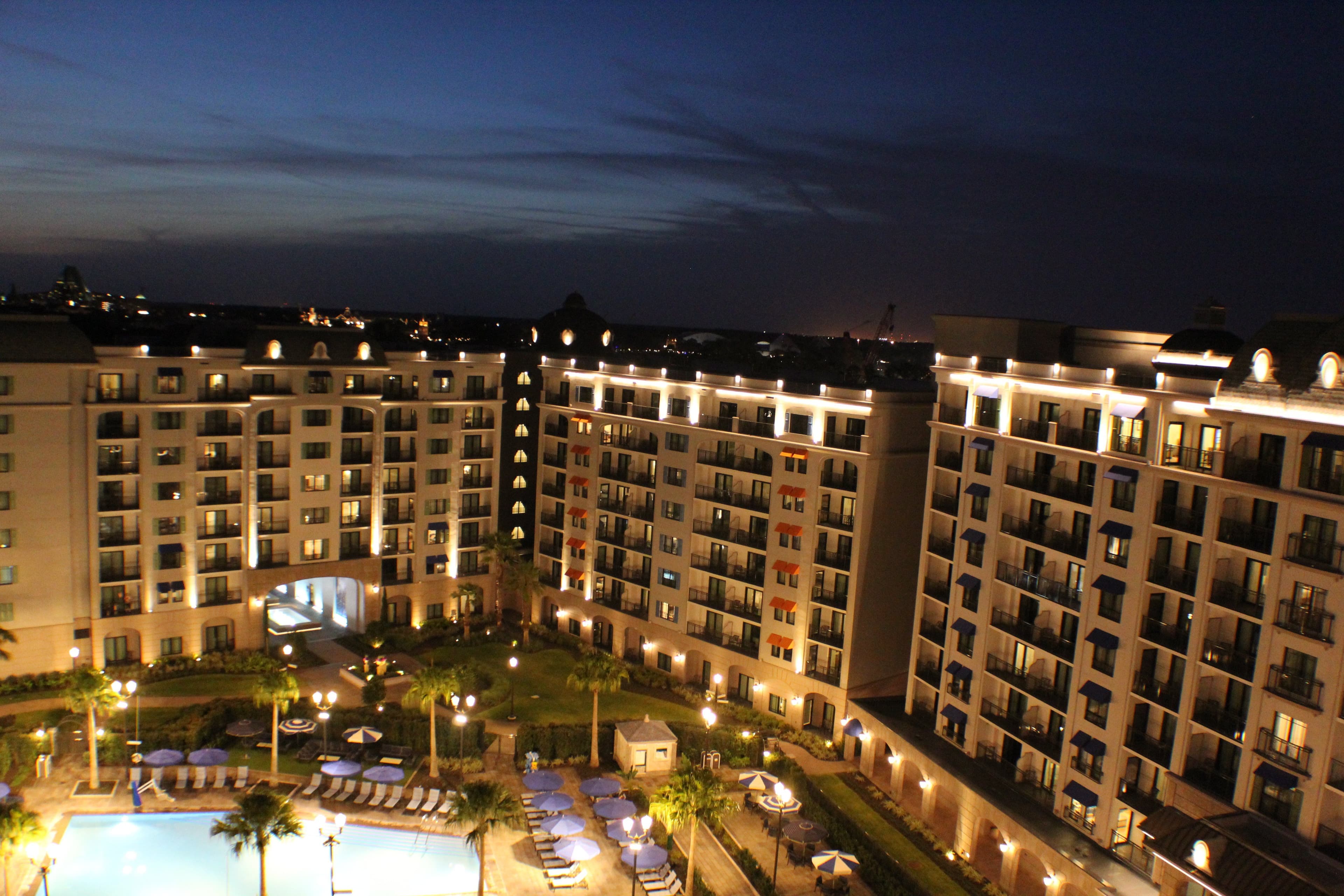 Resort hotel complex at dusk with illuminated multi-story buildings surrounding a central courtyard with pool and landscaped grounds.
