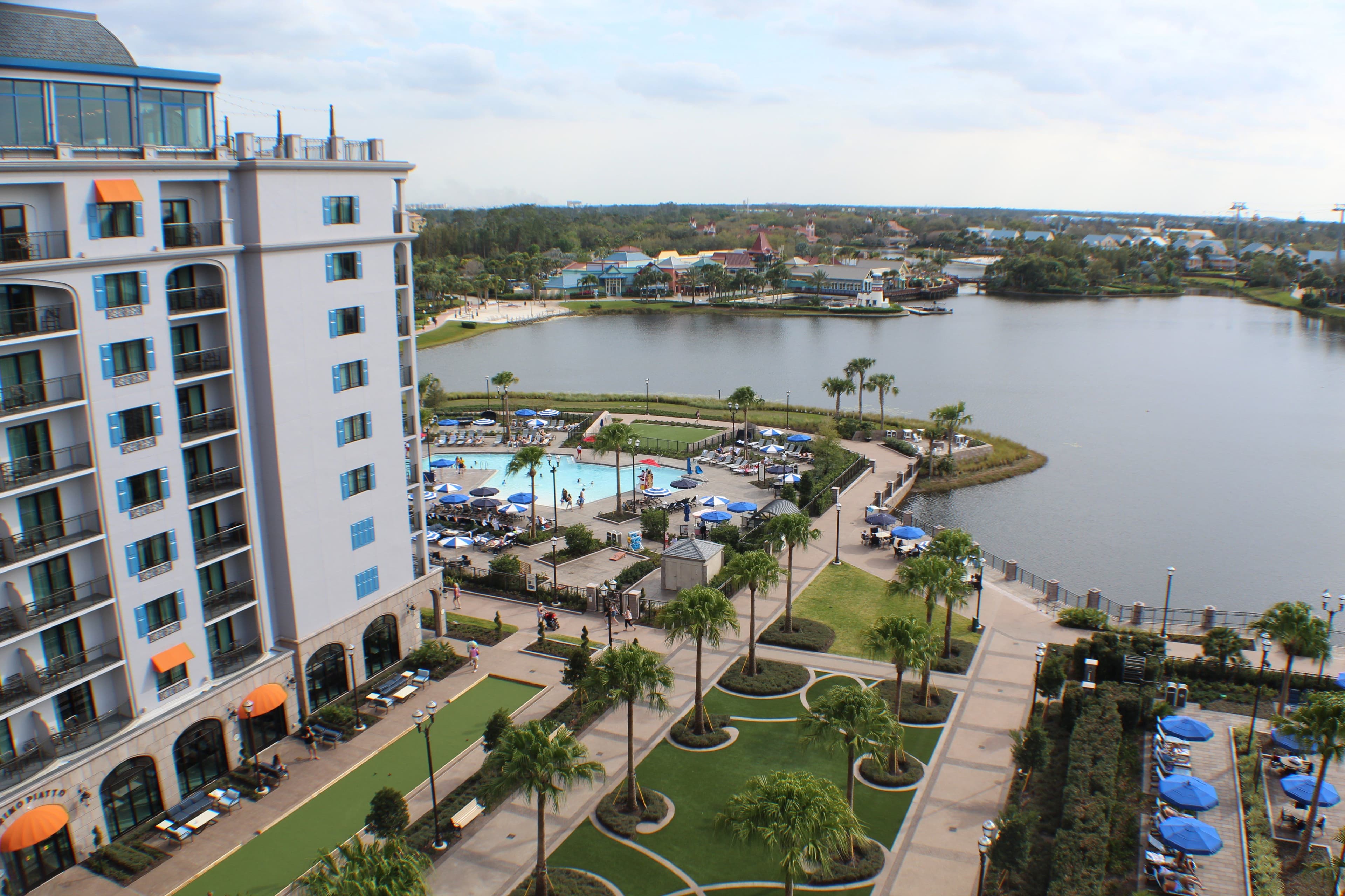 Aerial view of Disney resort with white hotel building, pool area, palm-lined walkways, and lakefront with boats in background