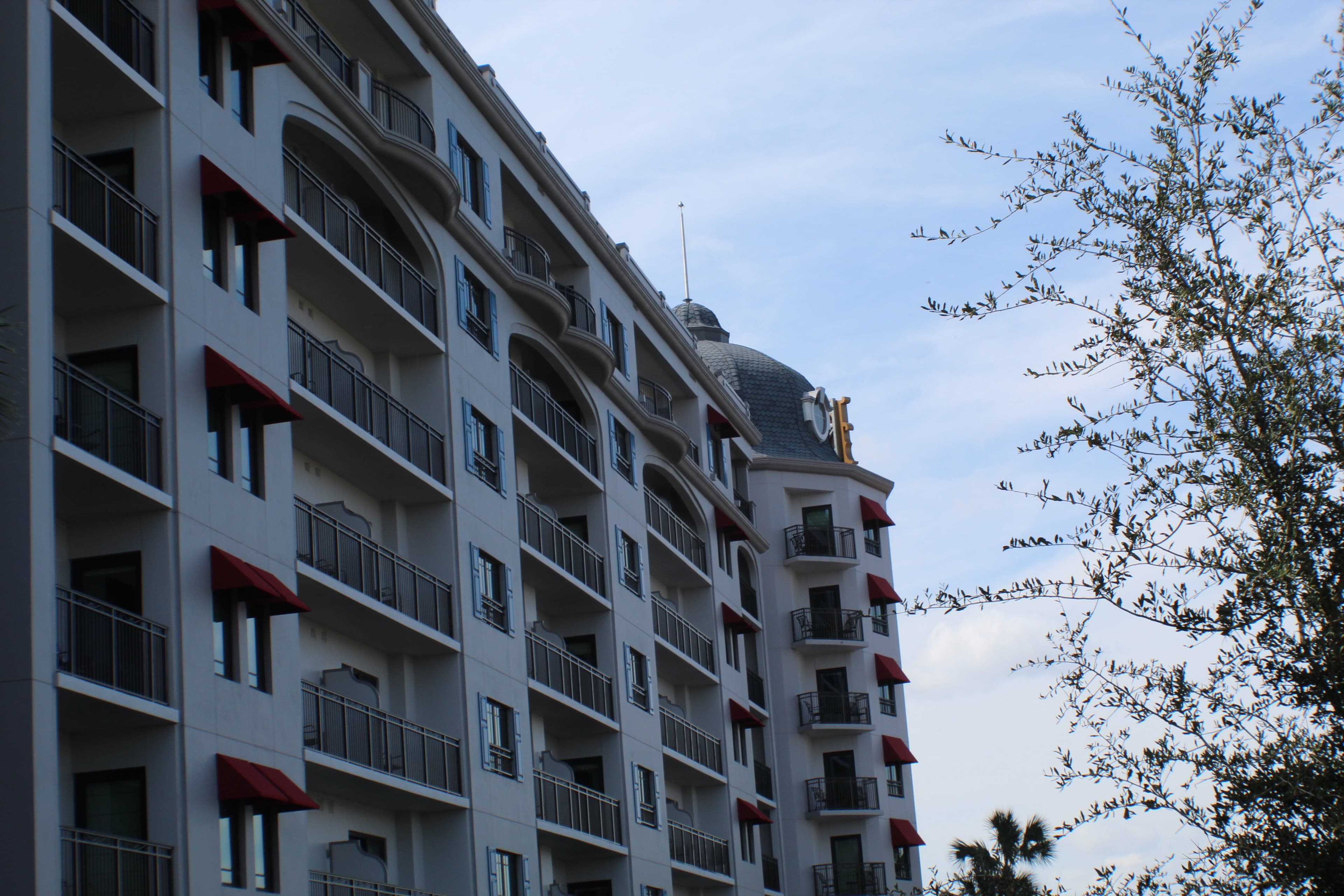 White multi-story resort building with red awnings and balconies under blue sky with trees visible on the right side.