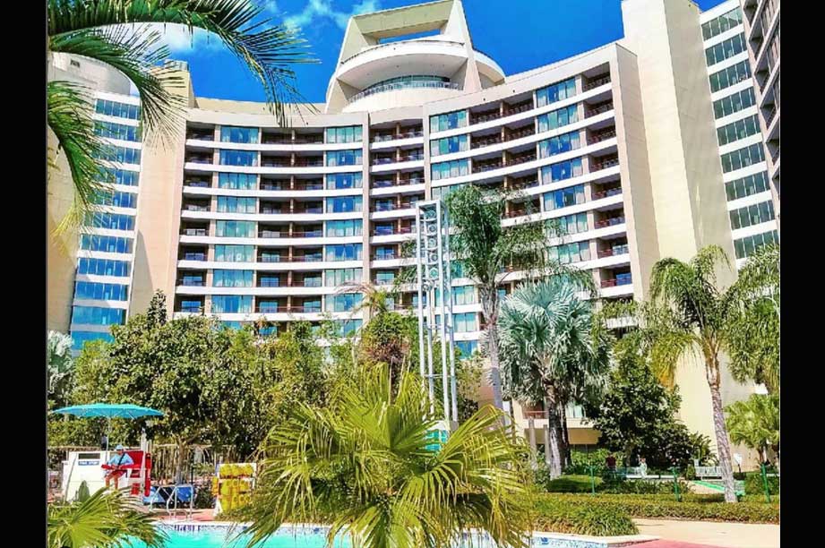 Modern curved resort building with multiple floors and balconies, surrounded by palm trees and tropical landscaping with a pool area visible in the foreground under blue sky.