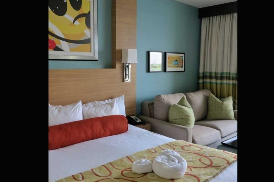 Modern hotel room with queen bed, gray sofa, blue accent wall, and colorful artwork in bright, contemporary style.