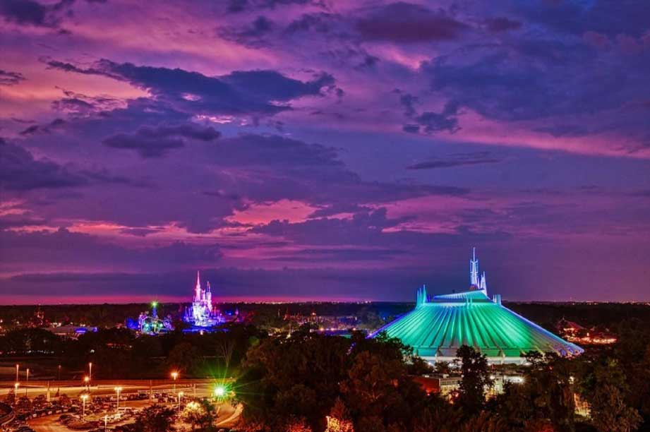 Stunning purple sunset sky over illuminated Disney World attractions including Space Mountain's green dome and Cinderella Castle in the distance.
