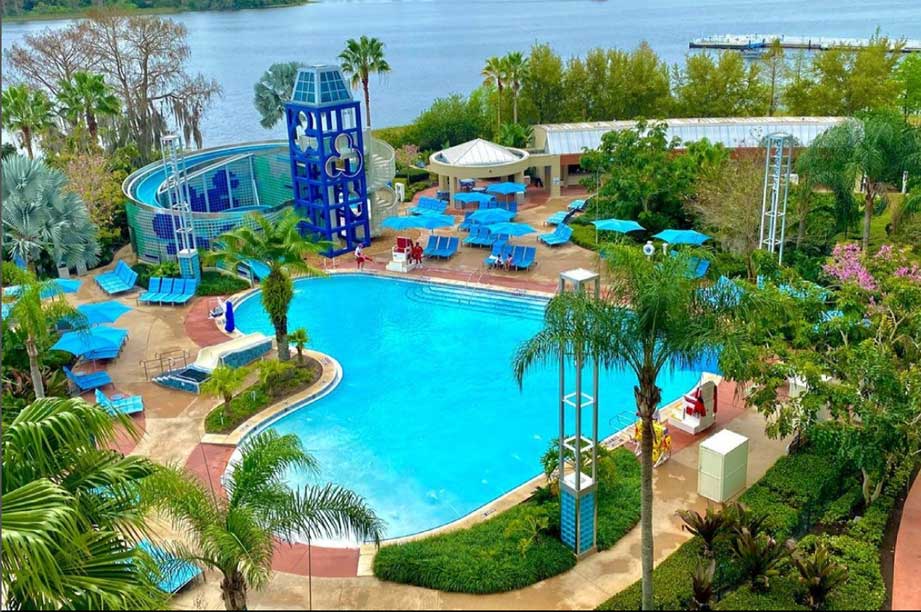 Resort pool area with water slide, blue umbrellas, palm trees, and waterfront view at tropical vacation destination.