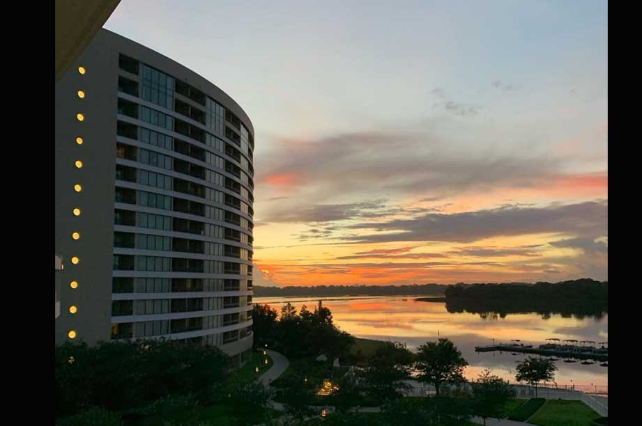 Modern curved resort tower overlooking a tranquil lake at sunset with orange and pink sky reflections on the water.