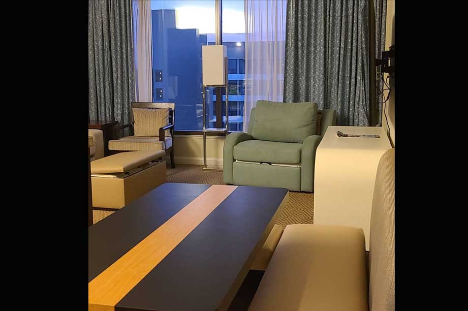 Modern hotel suite living area with green armchair, wooden furniture, floor lamp, and city view through large windows with gray curtains.
