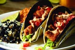 Colorful tacos filled with meat, lettuce and tomatoes served on a white plate with black beans and rice as sides
