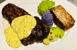 Gourmet plated dish featuring dark sausages, yellow scrambled eggs, purple potatoes, and fresh greens on white plate