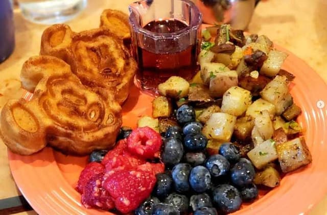 Orange plate with Mickey-shaped waffles, fresh berries, roasted potatoes, and syrup on wooden table at Disney resort.