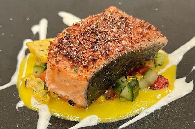 Seared salmon fillet with crispy skin on yellow sauce with diced vegetables and white cream drizzle on dark plate