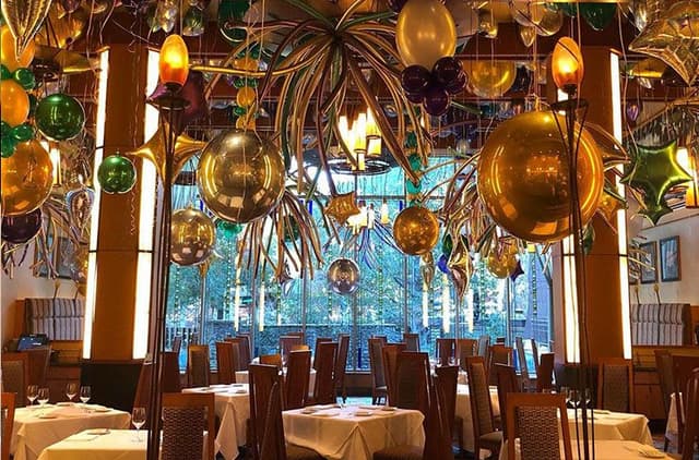 Elegant restaurant interior with decorative glass spheres and palm fronds hanging from ceiling, white-clothed tables, and large windows overlooking greenery.