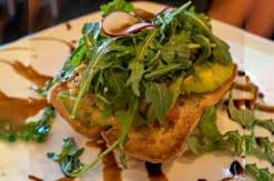 Grilled sandwich topped with fresh arugula and herbs on white plate at restaurant table