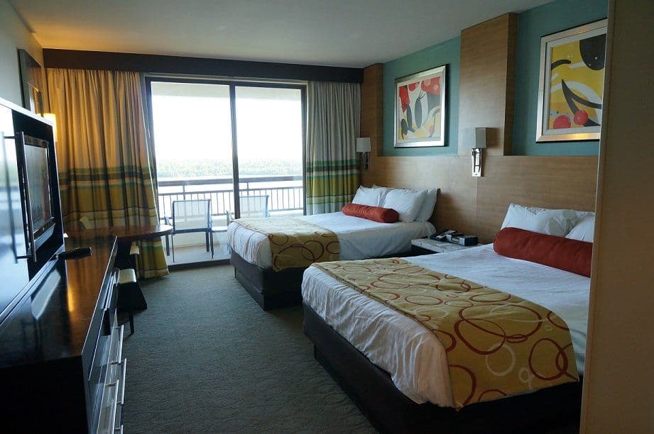 Modern hotel room with two beds, colorful bedding, large window with balcony view, flat-screen TV, and contemporary decor.