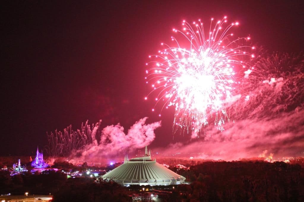 Spectacular red and pink fireworks exploding over Disney theme park at night with Space Mountain and Cinderella Castle visible below.