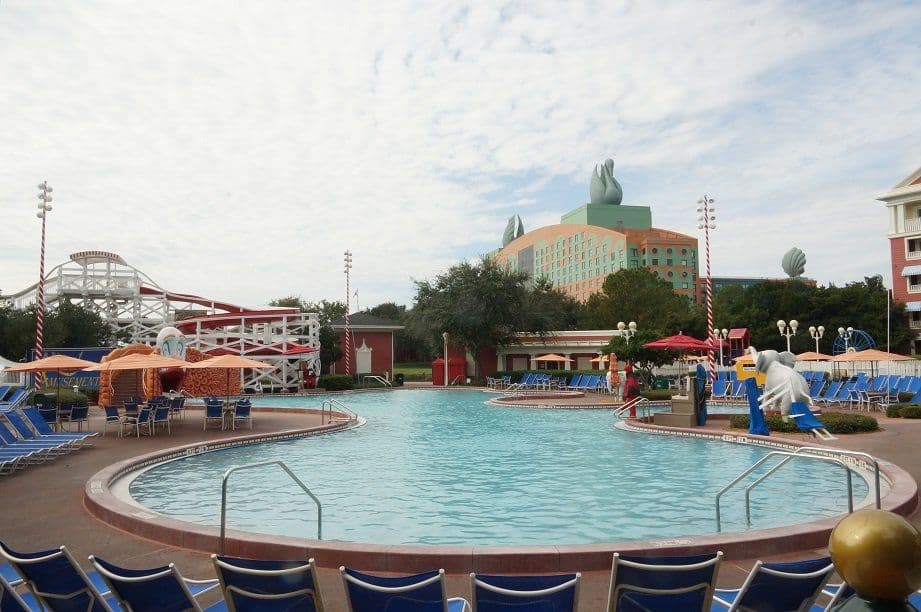 Large circular pool with blue lounge chairs, water slide, and Disney resort hotel with green spires in background under cloudy sky.