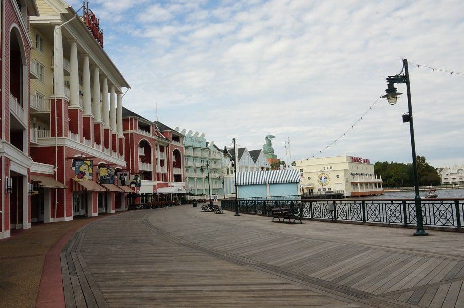 Colorful waterfront boardwalk with colonial-style buildings, white columns, and a cruise ship docked at the pier under cloudy skies.
