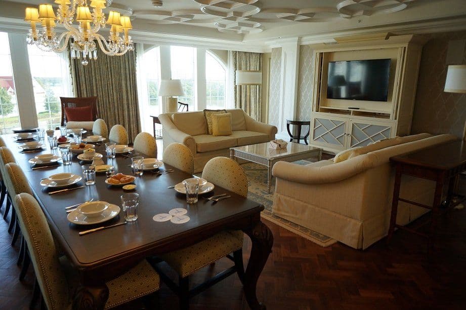 Luxurious resort suite with formal dining table, living area with beige sofas, crystal chandelier, and large windows overlooking scenic views.