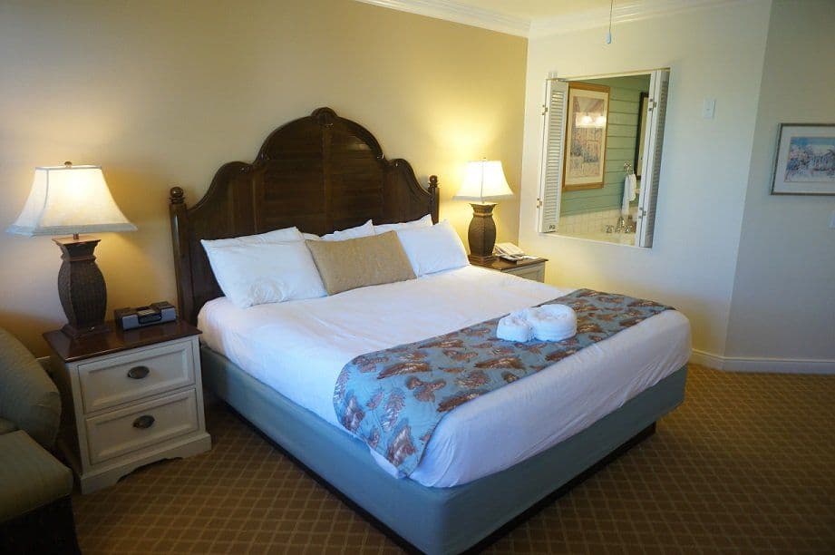 Hotel bedroom with king bed, dark wooden headboard, white nightstands, table lamps, and view into bathroom with green shutters.