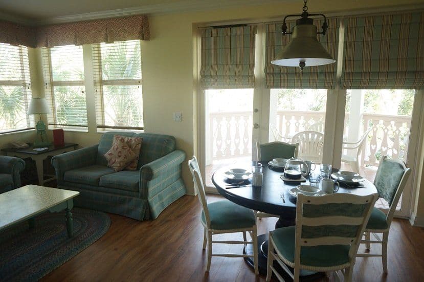 Bright dining area with round dark table, striped upholstered chairs, and adjoining living room featuring teal sofa and large windows with Roman shades.