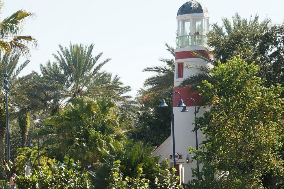 Colorful lighthouse with red and white stripes surrounded by lush tropical palm trees and greenery under a clear sky.