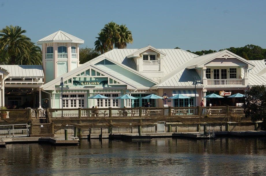 Waterfront Disney resort buildings with pastel colors, metal roofs, and wooden dock extending over calm water with palm trees