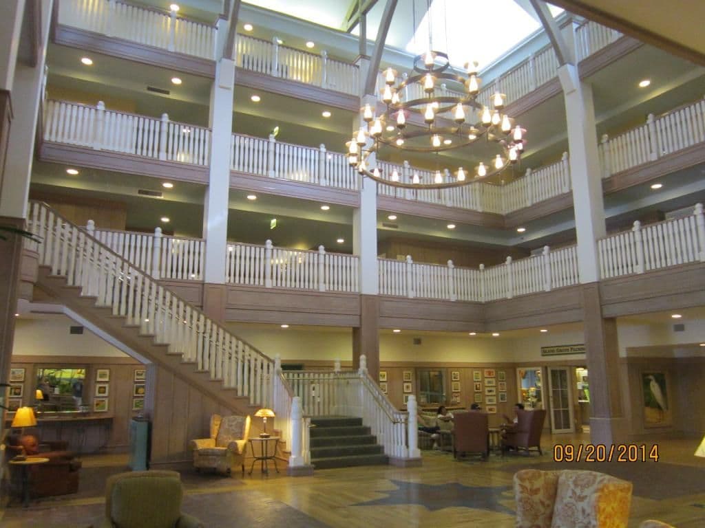 Grand hotel lobby with multi-story white balconies, ornate chandelier, curved staircase, and elegant seating areas under natural skylight