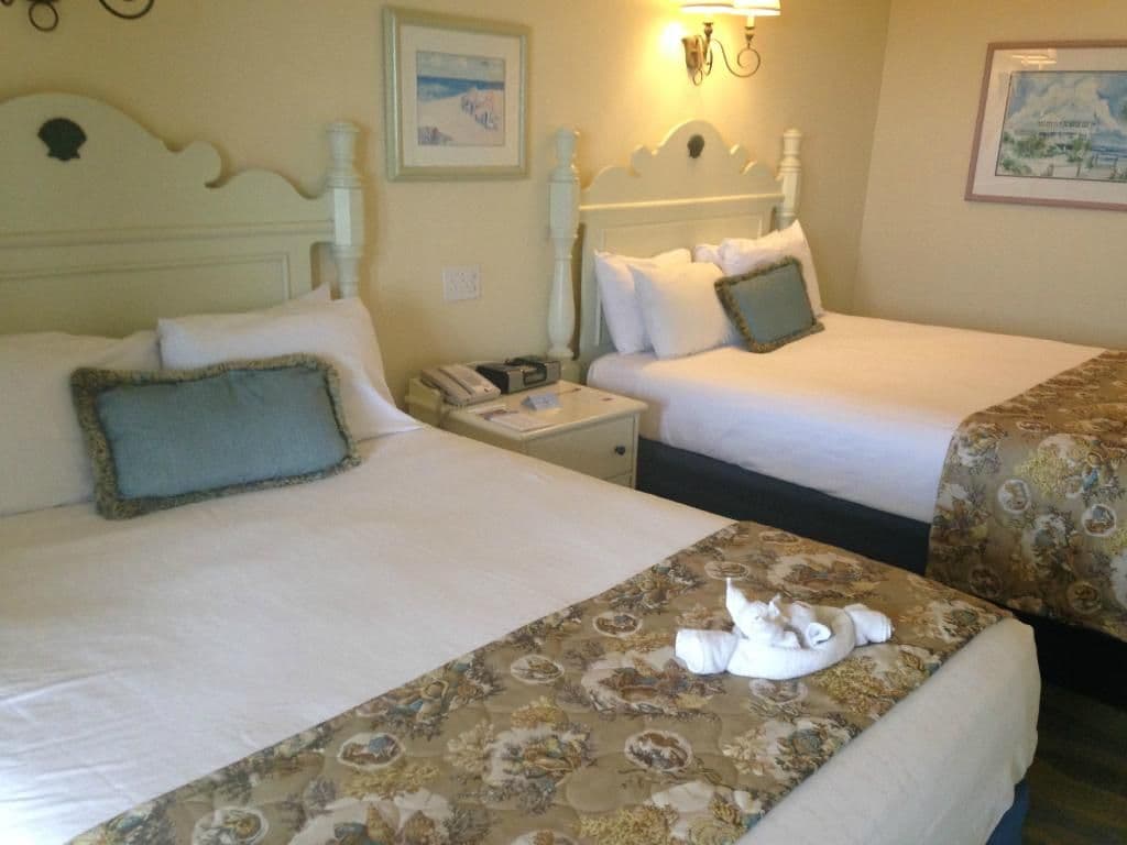 Two white beds with ornate golden bed runners and teal accent pillows in a hotel room with beach-themed artwork and warm lighting.
