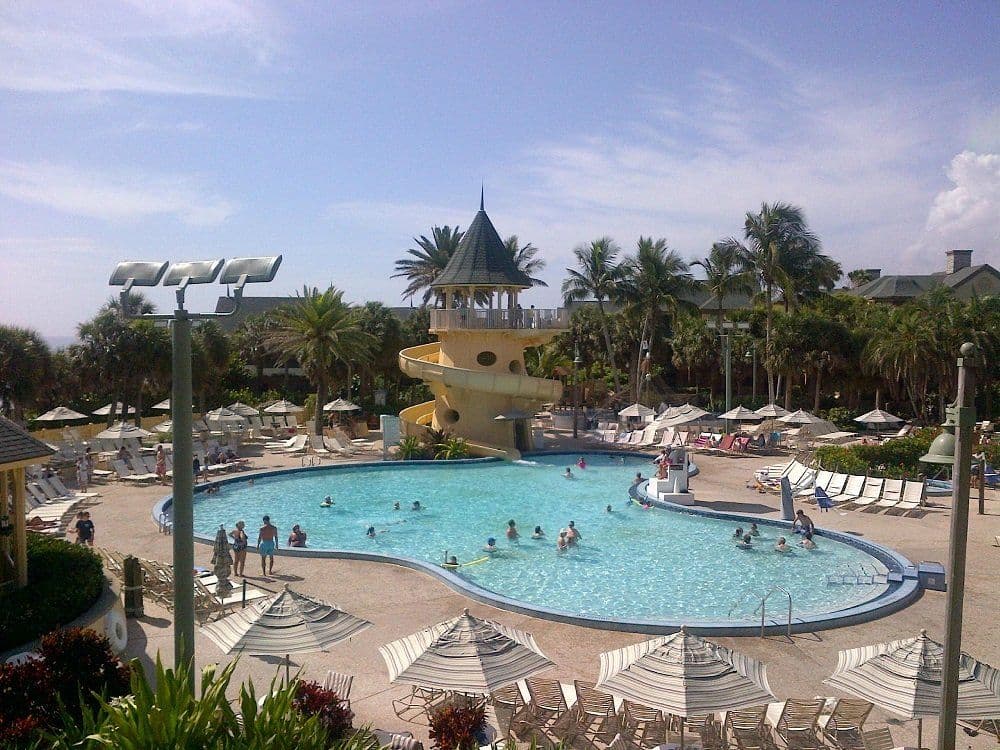 Large resort pool with yellow water slide and tower, surrounded by palm trees, lounge chairs, and umbrellas under blue sky.