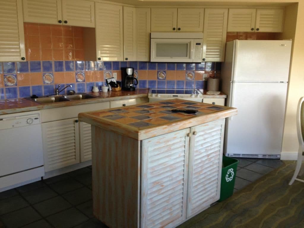 Kitchen with white louvered cabinets, colorful tile backsplash, small island with checkerboard countertop, and stainless steel appliances.