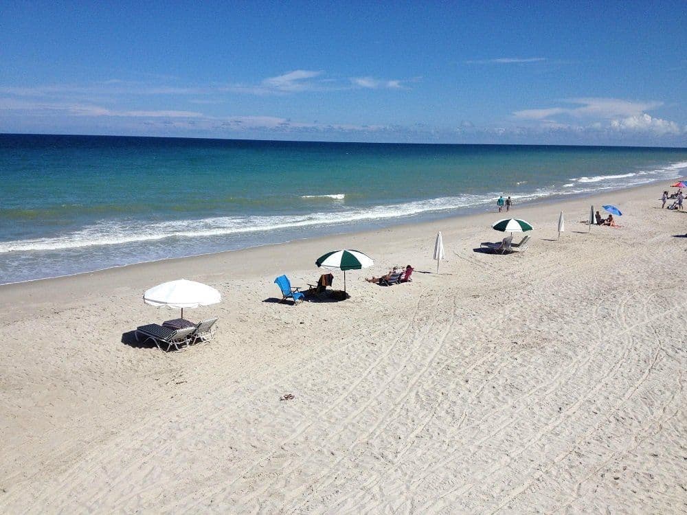Sandy beach with umbrellas, chairs, and beachgoers along turquoise ocean waters under blue sky with white clouds