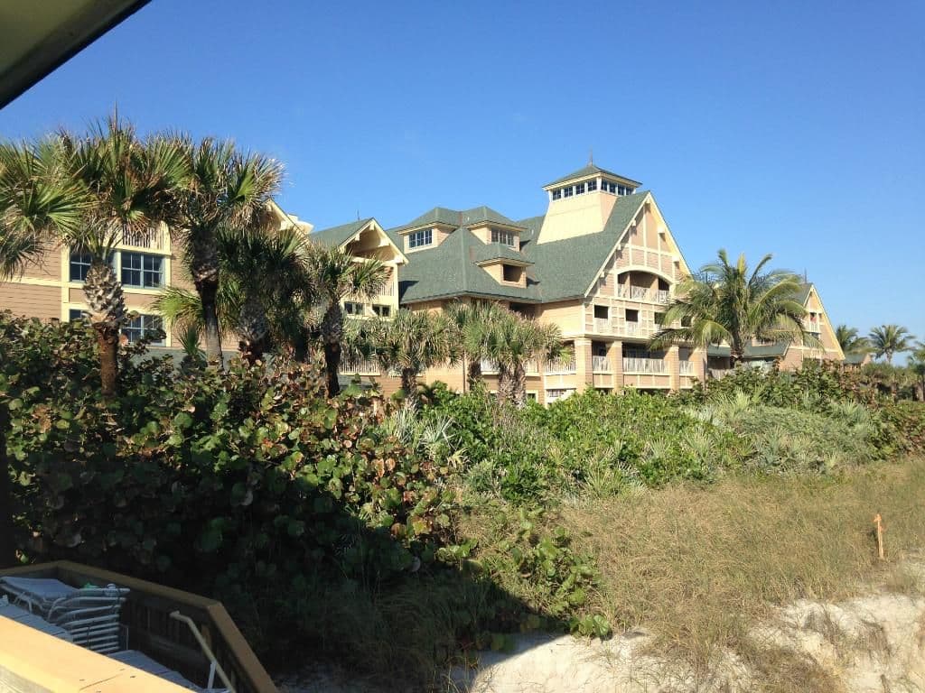 Beachfront Disney Vacation Club resort buildings with tropical landscaping, palm trees, and sandy dunes under blue sky